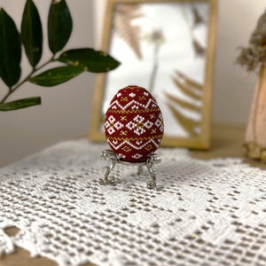 Pu&ograve; includere: Un uovo di Pasqua rosso, bianco e oro perlato con un motivo geometrico su un supporto argentato. L'uovo &egrave; su un tovagliolo bianco all'uncinetto.