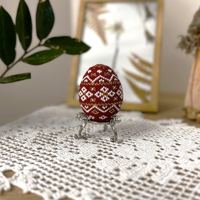 Pu&ograve; includere: Un uovo di Pasqua rosso, bianco e oro perlato con un motivo geometrico su un supporto argentato. L'uovo &egrave; su un tovagliolo bianco all'uncinetto.