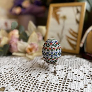 Peut inclure: Un &oelig;uf de P&acirc;ques perl&eacute; avec un motif g&eacute;om&eacute;trique dans des tons de bleu, vert, rouge et blanc. L'&oelig;uf est pr&eacute;sent&eacute; sur un support argent&eacute;.