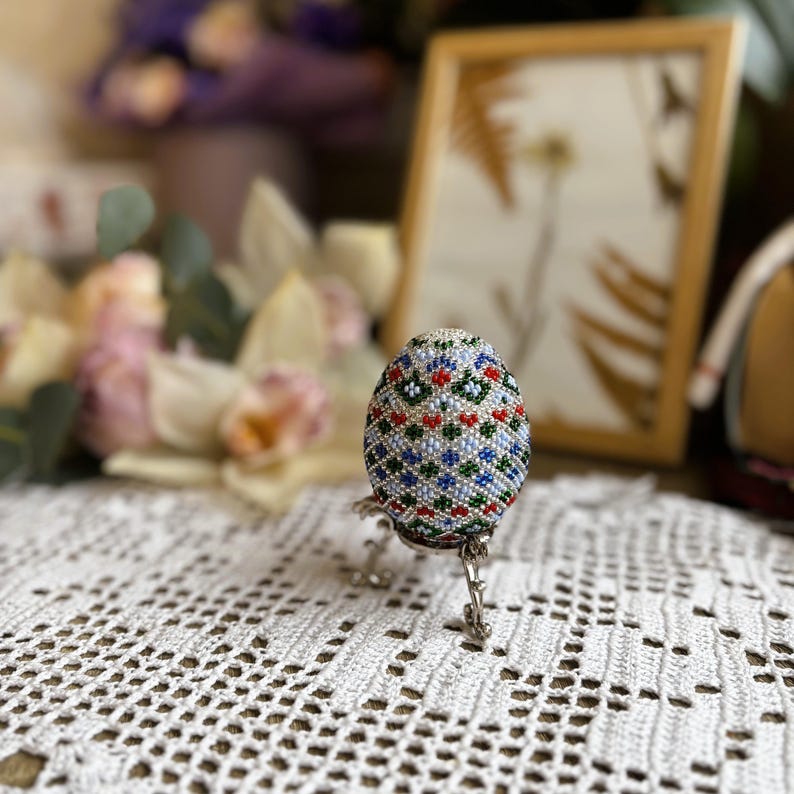 Peut inclure: Un &oelig;uf de P&acirc;ques perl&eacute; avec un motif g&eacute;om&eacute;trique dans des tons de bleu, vert, rouge et blanc. L'&oelig;uf est pr&eacute;sent&eacute; sur un support argent&eacute;.