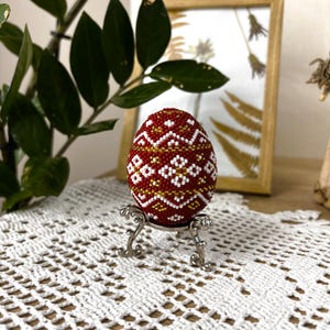 Pu&ograve; includere: Un uovo di Pasqua rosso, bianco e dorato perlato con un motivo geometrico su un supporto argentato. L'uovo &egrave; su una tovaglia all'uncinetto bianca.