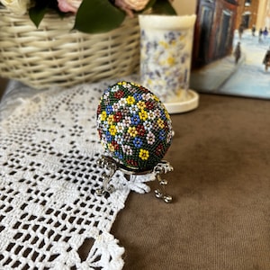 Peut inclure: Un &oelig;uf de P&acirc;ques perl&eacute; avec un motif floral dans des tons de rouge, jaune, bleu, vert et blanc. L'&oelig;uf est pr&eacute;sent&eacute; sur un support argent&eacute;.