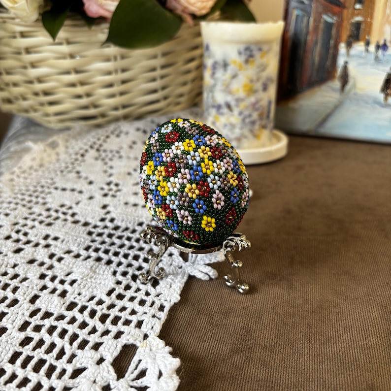 Peut inclure: Un &oelig;uf de P&acirc;ques perl&eacute; avec un motif floral dans des tons de rouge, jaune, bleu, vert et blanc. L'&oelig;uf est pr&eacute;sent&eacute; sur un support argent&eacute;.