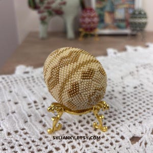 Puede incluir: Un huevo decorativo cubierto de cuentas de color crema con detalles dorados, exhibido sobre un soporte dorado ornamentado. El huevo presenta un patr&oacute;n geom&eacute;trico. El nombre de la tienda Etsy, SYLIANKY.ETSY.COM, es visible en la parte inferior.