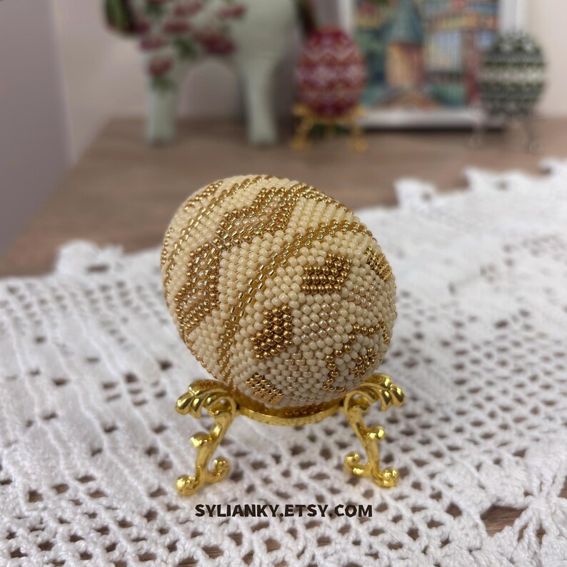 Puede incluir: Un huevo decorativo cubierto de cuentas de color crema con detalles dorados, exhibido sobre un soporte dorado ornamentado. El huevo presenta un patr&oacute;n geom&eacute;trico. El nombre de la tienda Etsy, SYLIANKY.ETSY.COM, es visible en la parte inferior.
