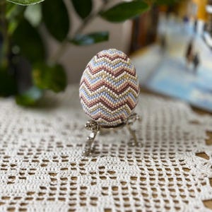 Peut inclure: Un &oelig;uf d&eacute;coratif avec un motif en chevrons perl&eacute; dans des tons de rose, bleu, or et blanc. L'&oelig;uf est pr&eacute;sent&eacute; sur un support en m&eacute;tal argent&eacute; avec un design d&eacute;taill&eacute;, pos&eacute; sur une nappe en dentelle blanche.