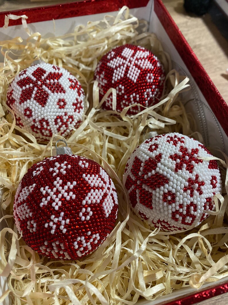 Op de afbeelding: Vier rode en witte kerstballen van kralen met sneeuwvlokmotieven in een doos gevuld met gescheurd papier.