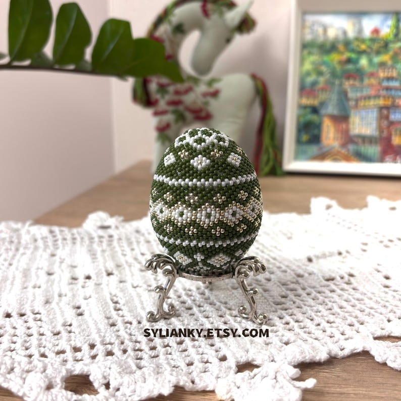 Może przedstawiać: Oliwkowe jajko z koralik&oacute;w z białymi i srebrnymi akcentami, prezentowane na srebrnym stojaku. Jajko ma geometryczny wz&oacute;r i jest umieszczone na białym koronkowej obrusie i obrazie. Widoczna jest nazwa sklepu Etsy, SYLIANKY.ETSY.COM.