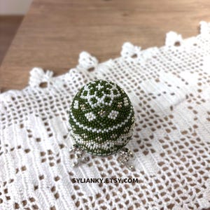 Może przedstawiać: Ozdobne jajko z koralik&oacute;w z zielono-białym kwiatowym wzorem. Jajko jest ozdobione małymi koralikami i ma srebrny metaliczny frędzel. Widoczny jest tekst "SYLIANKY.ETSY.COM".
