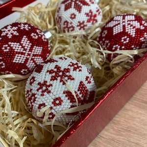 Op de afbeelding: Vier rode en witte kerstballen van kralen met sneeuwvlokmotieven in een rode doos met gescheurd papier.