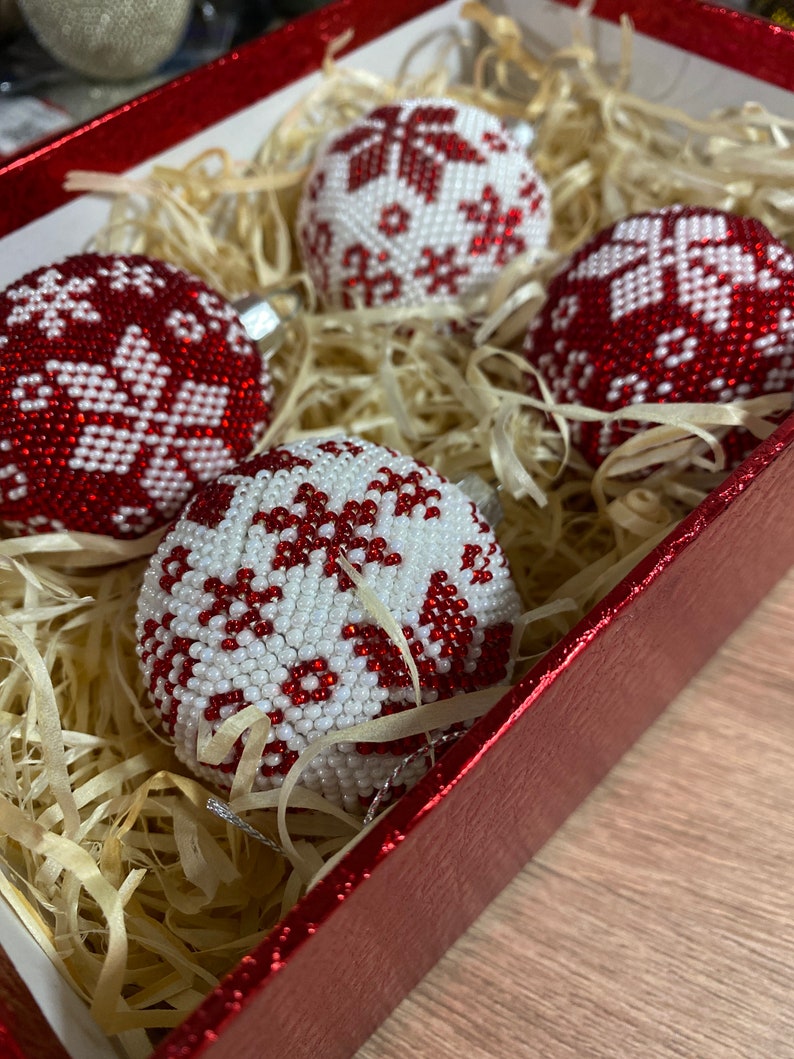 Op de afbeelding: Vier rode en witte kerstballen van kralen met sneeuwvlokmotieven in een rode doos met gescheurd papier.