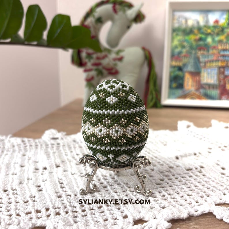 Może przedstawiać: Ozdobne, zielono-białe jajko z koralik&oacute;w stoi na srebrnym metalowym stojaku na białej koronkowej serwetce. Jajko ma geometryczny wz&oacute;r. Nazwa sklepu Etsy, SYLIANKY.ETSY.COM, jest widoczna na dole zdjęcia.
