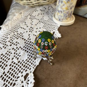 Peut inclure: Un &oelig;uf de P&acirc;ques vert d&eacute;cor&eacute; de perles color&eacute;es dans un motif floral, pr&eacute;sent&eacute; sur un support argent&eacute;.