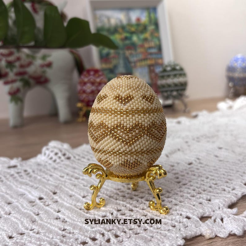 Puede incluir: Un huevo decorativo cubierto de cuentas de color crema con detalles dorados, exhibido sobre un soporte dorado ornamentado. El huevo presenta un patr&oacute;n geom&eacute;trico. El nombre de la tienda Etsy, SYLIANKY.ETSY.COM, es visible en la parte inferior.