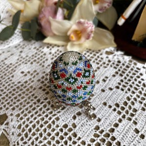Peut inclure: Un &oelig;uf de P&acirc;ques perl&eacute; avec un fond blanc et un motif floral color&eacute; en rouge, bleu et vert. L'&oelig;uf est sur un support argent&eacute;.