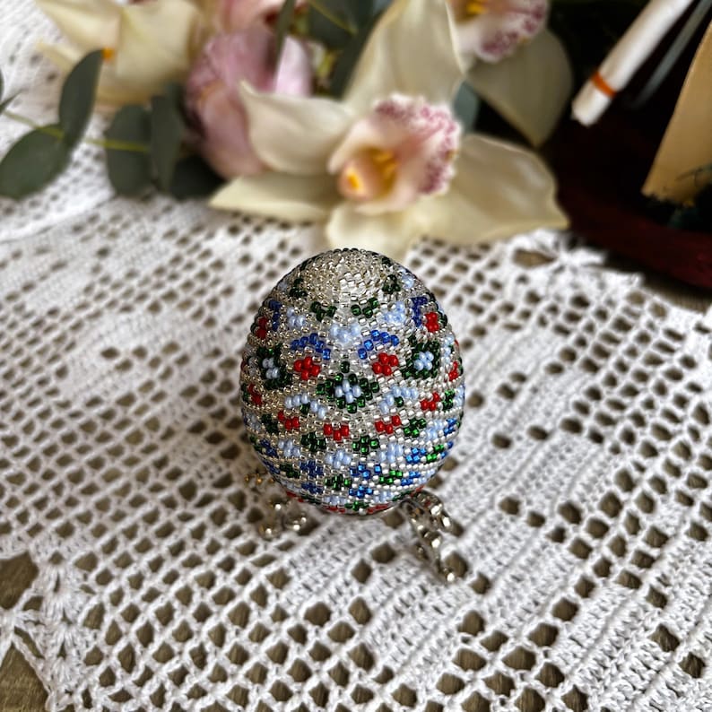 Peut inclure: Un &oelig;uf de P&acirc;ques perl&eacute; avec un fond blanc et un motif floral color&eacute; en rouge, bleu et vert. L'&oelig;uf est sur un support argent&eacute;.