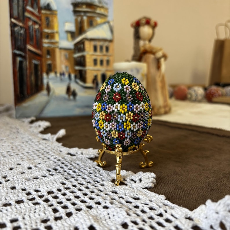 Peut inclure: Un &oelig;uf de P&acirc;ques vert d&eacute;cor&eacute; de fleurs color&eacute;es est pr&eacute;sent&eacute; sur un support dor&eacute;. L'&oelig;uf est fait de perles et est une pysanka ukrainienne traditionnelle.