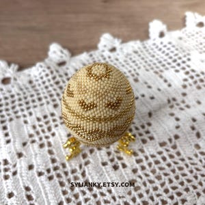 Puede incluir: Un huevo decorativo cubierto de peque&ntilde;as cuentas de color crema con detalles dorados. El huevo est&aacute; adornado con patrones de cuentas doradas y descansa sobre un soporte dorado. El nombre de la tienda Etsy, SYLIANKY.ETSY.COM, es visible en la parte inferior.