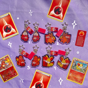 May include: Six clear acrylic keychains featuring Pokemon characters from the Fire type. Each keychain has a red and white Pocky snack wrapper design. The keychains are arranged in two rows of three. The keychains are on a purple background with red and white energy cards and a Fennekin card.