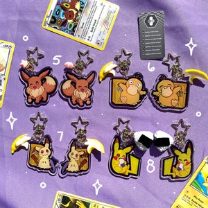 May include: Eight clear acrylic keychains featuring different Pokemon characters. The keychains are on a purple background with Pokemon trading cards scattered around them. The characters include Eevee, Pikachu, Pichu, and Psyduck.
