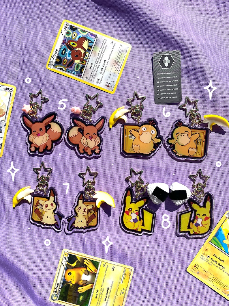 May include: Eight clear acrylic keychains featuring different Pokemon characters. The keychains are on a purple background with Pokemon trading cards scattered around them. The characters include Eevee, Pikachu, Pichu, and Psyduck.