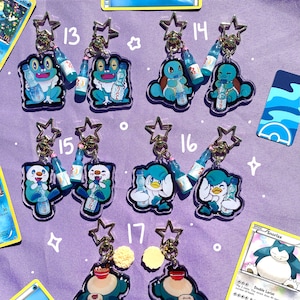 May include: A set of 10 clear acrylic keychains featuring different Pokemon characters drinking a blue beverage. Each keychain has a gold star charm and a gold ring.