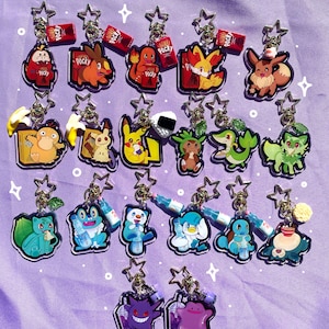 May include: A collection of 18 colorful acrylic charms featuring Pokemon characters. The charms are attached to gold keychains and feature various Pokemon, including Pikachu, Bulbasaur, Squirtle, Charmander, Eevee, and more. The charms are displayed on a purple background.