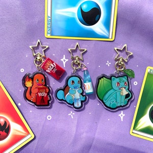May include: Three clear acrylic keychains featuring Pokemon characters: Charmander, Squirtle, and Bulbasaur. Each keychain has a different snack: Charmander has a box of Pocky, Squirtle has a bottle of Ramune, and Bulbasaur has a green leaf.
