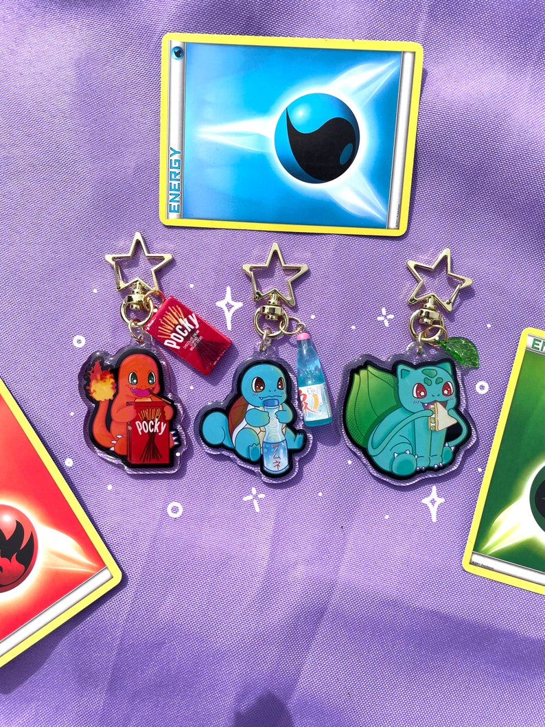 May include: Three clear acrylic keychains featuring Pokemon characters: Charmander, Squirtle, and Bulbasaur. Each keychain has a different snack: Charmander has a box of Pocky, Squirtle has a bottle of Ramune, and Bulbasaur has a green leaf.