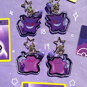 May include: Four purple and white Pokemon character charms with boba tea. The charms are attached to gold chains with star-shaped charms. The characters are Gengar and Jigglypuff.