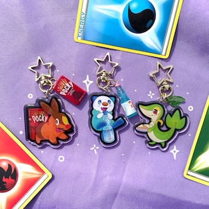 May include: Three colorful anime-style character keychains with gold star-shaped charms. The characters are holding snacks, including a green character holding a plate of food, a blue character holding a bottle of soda, and an orange character holding a box of Pocky.