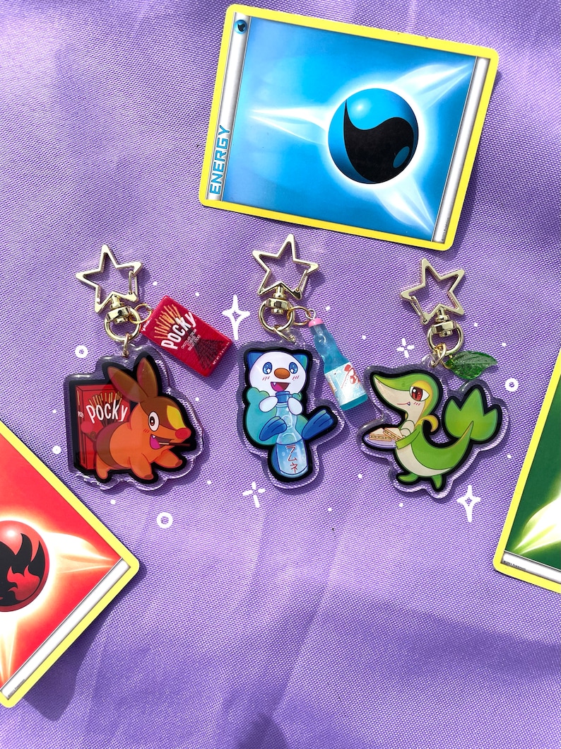 May include: Three colorful anime-style character keychains with gold star-shaped charms. The characters are holding snacks, including a green character holding a plate of food, a blue character holding a bottle of soda, and an orange character holding a box of Pocky.