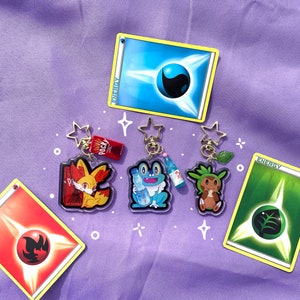 May include: Four Pokemon character keychains with a red, blue, green, and yellow energy card. The keychains are made of clear acrylic and feature the characters Vulpix, Froakie, Grookey, and a water bottle. The energy cards are from the Pokemon Trading Card Game.