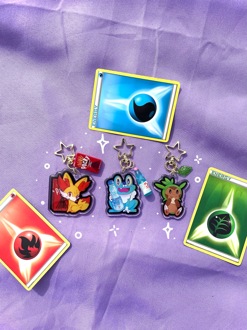 May include: Four Pokemon character keychains with a red, blue, green, and yellow energy card. The keychains are made of clear acrylic and feature the characters Vulpix, Froakie, Grookey, and a water bottle. The energy cards are from the Pokemon Trading Card Game.