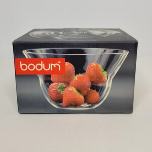 May include: A box featuring a clear glass bowl filled with fresh strawberries. The bowl is labeled "bodum" in red. The strawberries are bright red with green leaves, set against a dark background.