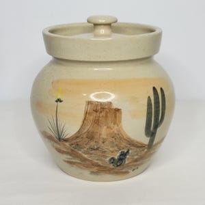 May include: A beige ceramic jar with a lid, featuring a hand-painted desert landscape. The design includes a mesa, cactus, and other desert plants in shades of brown, green, and yellow. The jar has a rounded shape and a small knob on the lid.
