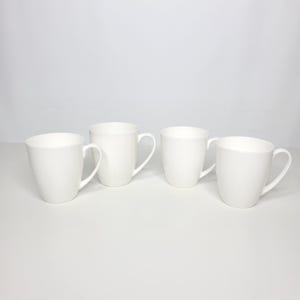 May include: A set of four plain white ceramic mugs arranged in a row. Each mug features a smooth, glossy finish, a rounded body, and a curved handle on the side. The mugs are uniform in color and design, presenting a clean, simple aesthetic.