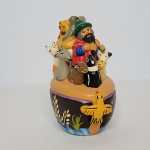 May include: A ceramic figurine of a man with a beard and a hat, holding a staff and surrounded by animals. The figurine is painted in bright colors and has a whimsical style. The man is standing on a boat with a yellow bird on the side.