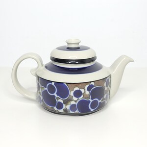 May include: A ceramic teapot featuring a cream-colored body with a dark blue band around the lid and upper section. The lower portion is decorated with a repeating pattern of dark blue circular shapes and white flower outlines on a brown background.