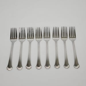 May include: Eight silver forks arranged in a row. Each fork has a long, slender handle with a decorative, scalloped base. The tines are evenly spaced and pointed. The forks are set against a plain, light background.