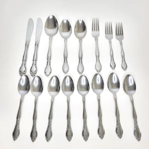 May include: A collection of stainless steel silverware, including spoons, forks, and knives, arranged on a white surface. The silverware has ornate handle detailing, reflecting light. The set includes various sizes of spoons, forks, and knives.