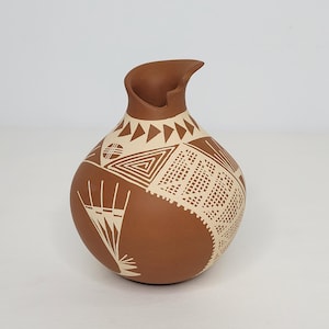 May include: A brown ceramic vase with a unique spout and a rounded body. The vase is decorated with geometric patterns in a cream color, including triangles, squares, and lines. The vase is likely handmade and suitable for home decor.