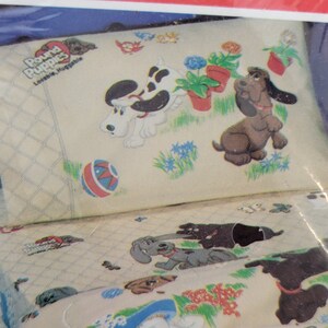 Pode incluir: Um tecido de cor creme com uma ilustra&ccedil;&atilde;o colorida de c&atilde;es brincando em um jardim. O tecido apresenta o texto "Pound Puppies" e "Lovable, Huggable".