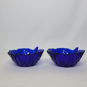 May include: Two cobalt blue glass bowls with a wavy, ruffled edge. The bowls are empty and sitting on a white surface.