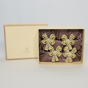 May include: A set of four gold-colored bow-shaped napkin rings presented in a cream-colored box. The napkin rings have a detailed, openwork design and are displayed on a brown satin fabric. The box is open, revealing the contents.