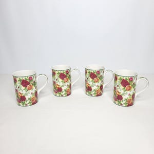 May include: A set of four white ceramic mugs, each featuring a repeating pattern of red and yellow roses with green leaves and stems. The mugs have a cylindrical shape, a curved handle on the side, and a thin green rim around the top edge.