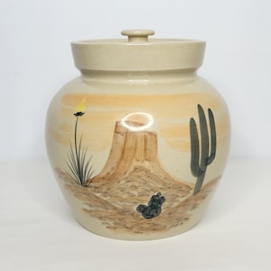 May include: A beige ceramic jar with a lid, hand-painted with a desert landscape scene. The design includes a mesa, cactus, and other desert plants. The jar is a round shape and has a small knob on the lid.