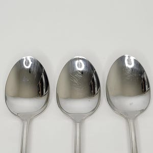 May include: Three stainless steel spoons on a white background. The spoons have oval bowls and long, slender handles. The spoons reflect light, appearing bright and shiny. These spoons are a common kitchen utensil, used for eating and serving food.