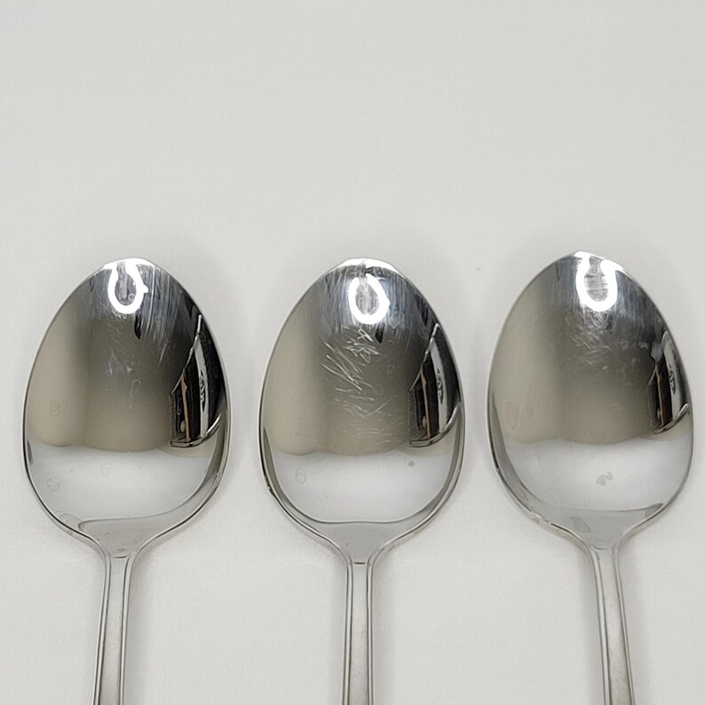 May include: Three stainless steel spoons on a white background. The spoons have oval bowls and long, slender handles. The spoons reflect light, appearing bright and shiny. These spoons are a common kitchen utensil, used for eating and serving food.