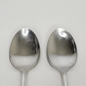 May include: Two stainless steel spoons with oval bowls and long, slender handles. The spoons are positioned side-by-side against a plain white background. The spoons reflect light, showing a shiny, metallic surface.
